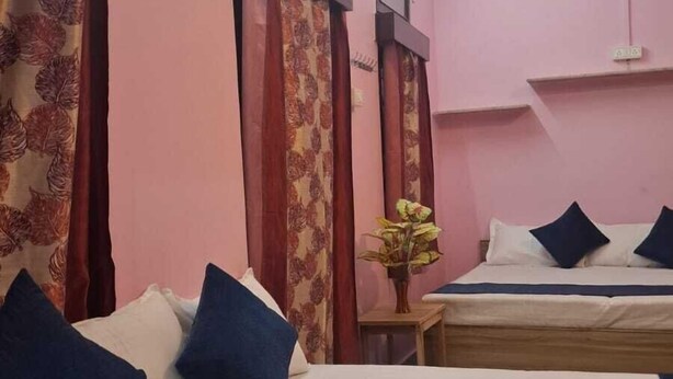 A hotel room with two beds, pink walls, floral curtains, a ceiling fan, and a small table with a plant. - Madhavi Homestay (Varanasi)