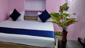 A bed with a white and blue bedspread, two dark pillows, and a wooden headboard. A potted plant with green leaves is visible to the right. - Madhavi Homestay (Varanasi)