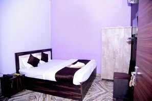 A neatly made bed with a wooden frame and dark bedding, a small bedside table with a phone and a book, and a wardrobe with a mirror. - Hotel Shree Mahalaxmi (Karchana)