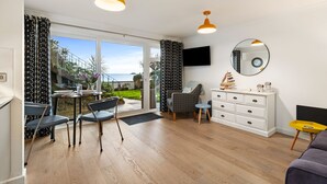 Un vaste espace de vie comprenant une grande porte-fenêtre coulissante donnant sur un balcon, une table à manger avec des chaises, un canapé, une commode blanche et un téléviseur fixé au mur.
