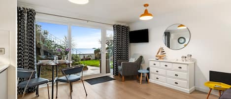 A spacious living area with a large sliding glass door leading to a balcony, a dining table with chairs, a sofa, a white dresser, and a television mounted on the wall.