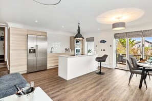 A modern kitchen with a central island, stainless steel appliances, and a dining area with a table and chairs. - Blue Horizon - Spectacular Coastal Views (Tenby)
