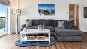 En moderne stue med en grå hjørnesofa, et hvidt sofabord, fletkurve og et stort vindue med udsigt til det fri.
