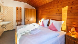 Ein Schlafzimmer mit Holzbalkendecke, einem Bett mit weißer Bettwäsche und roten Kissen, einem Holzschrank, einem Nachttisch mit Lampe sowie einem Badezimmer mit Waschbecken und gemustertem Wandpapier.