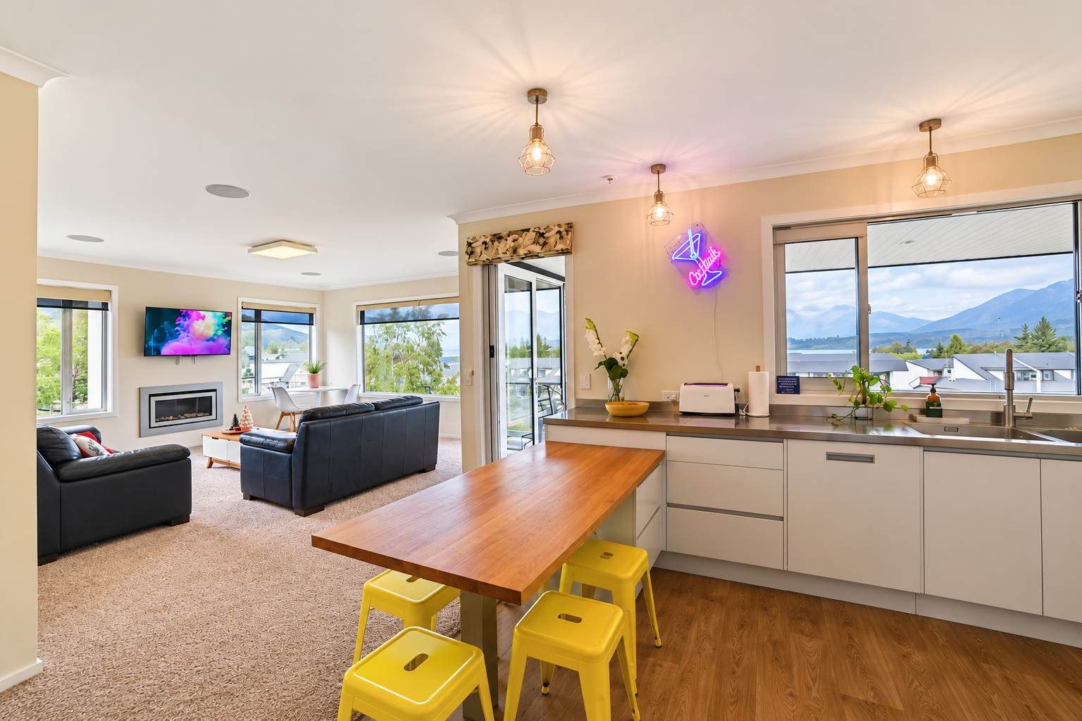 A modern kitchen with a wooden table and yellow stools, a flat-screen TV, and a neon sign.