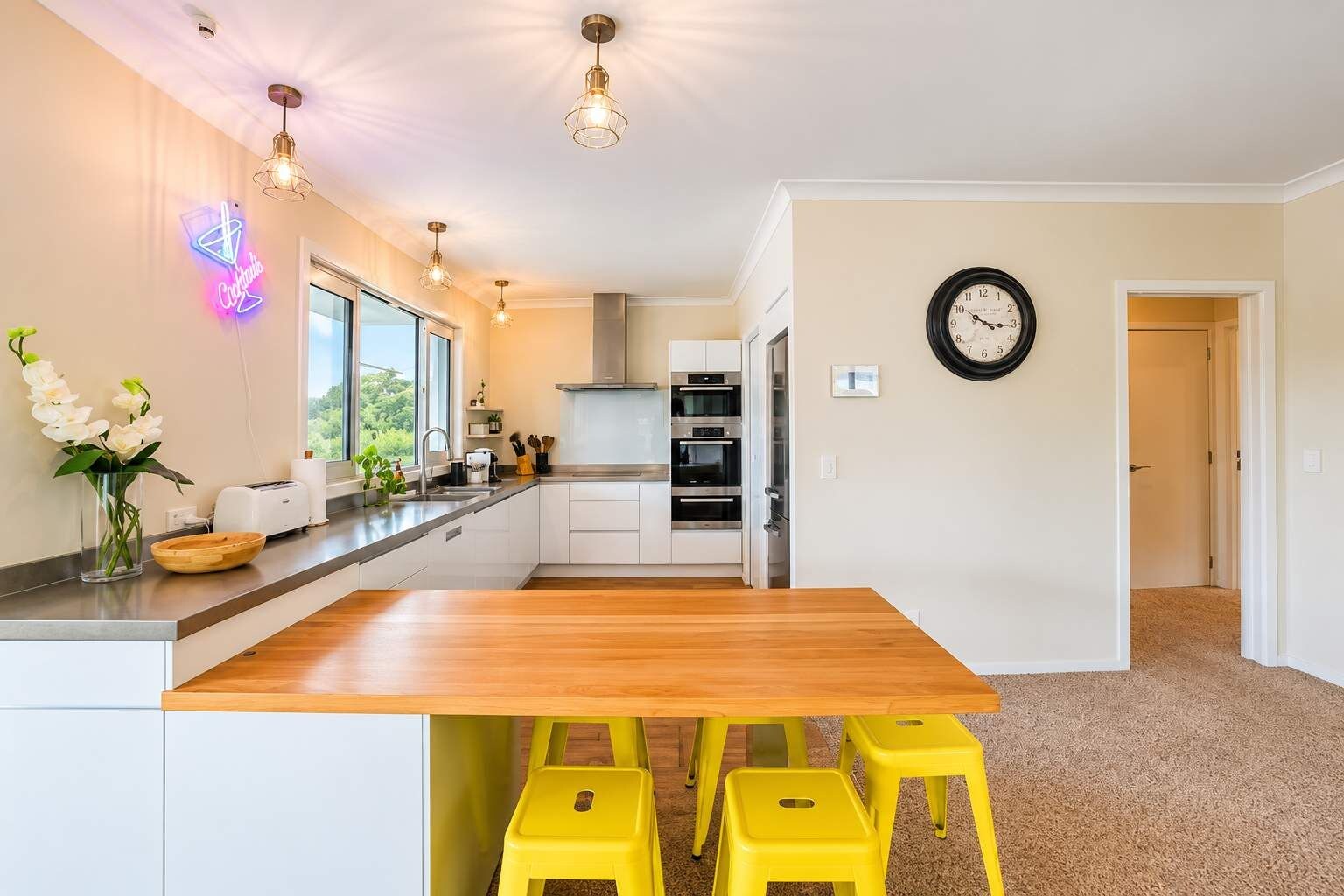 A modern kitchen with a wooden island, yellow stools, a neon sign, and a clock on the wall.