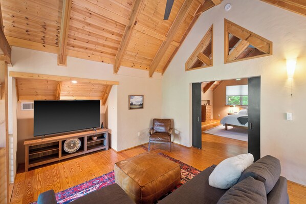 A spacious living room with a wooden ceiling, a large flat-screen TV on a wooden stand, a leather ottoman, and a brown sofa with a white pillow. - Private Mountain Retreat near Breweries and Wineries (Lovingston)