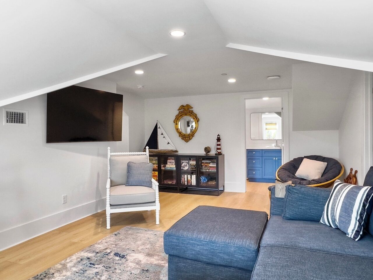 A spacious loft with a high ceiling, a large flat-screen TV mounted on the wall, a dark wood cabinet with books, a white upholstered chair, and a blue sofa with striped pillows.