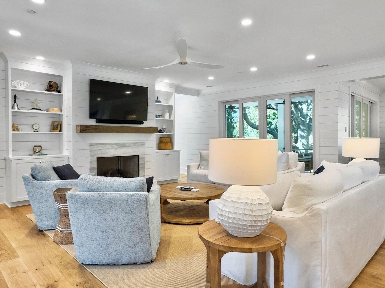 A modern living room with a fireplace, white sofas, a television, and a large window with a view of trees.