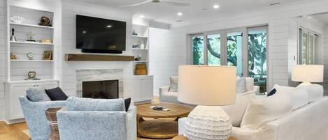 A modern living room with a fireplace, white sofas, a television, and a large window with a view of trees.