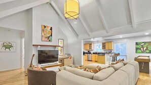 A modern living room with a large sectional sofa, a flat-screen TV, and a dining area with wooden cabinets. - 4 BR Beach House w/ Lush Backyard | Pet Friendly (Isle of Palms)