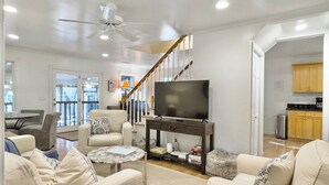A spacious living room with a large TV, comfortable seating, a staircase, and a view of the outdoors. - 4 BR Beach House w/ Lush Backyard | Pet Friendly (Isle of Palms)