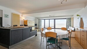 Eine moderne Küche mit einer zentralen Kochinsel, einem Essbereich mit rundem Tisch und Stühlen sowie einem großen Fenster mit Ausblick.