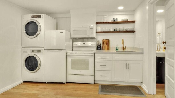 A compact kitchen with a washing machine, microwave, stove, and a sink with wooden shelves above. - Charming Historic Studio Cottage (Lakeland)