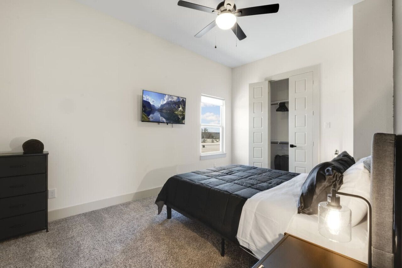 A bedroom with a bed, a dresser, a ceiling fan, a TV, and a window.