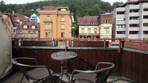 Un balcon avec deux chaises et une table, d’où l’on peut admirer un paysage urbain composé de différents bâtiments et, au loin, une colline boisée.