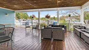 A covered outdoor area with wooden flooring, wicker furniture, and a view of boats and greenery. - Sandy Feet Retreat: Like-New, Harbor Club South Haven, Year-Round Pool & Hot Tub (South Haven)