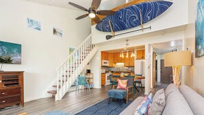 A modern living room with a staircase, a kitchen in the background, and a ceiling fan.