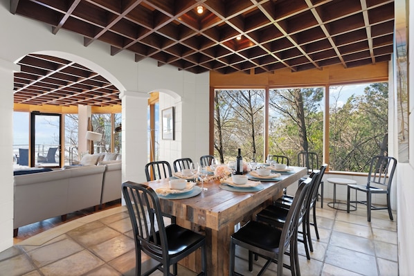 A dining area with a wooden table set for six, surrounded by black chairs, with a view of trees through large windows. - The Iconic Round House (St Simons Island)