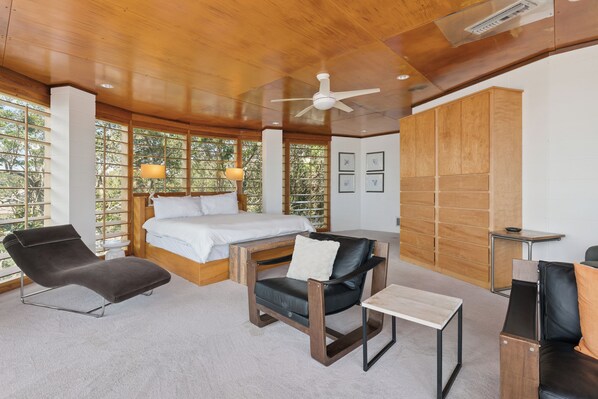 A bedroom with a bed, a desk, two chairs, a ceiling fan, and large windows with a view of trees. - The Iconic Round House (St Simons Island)