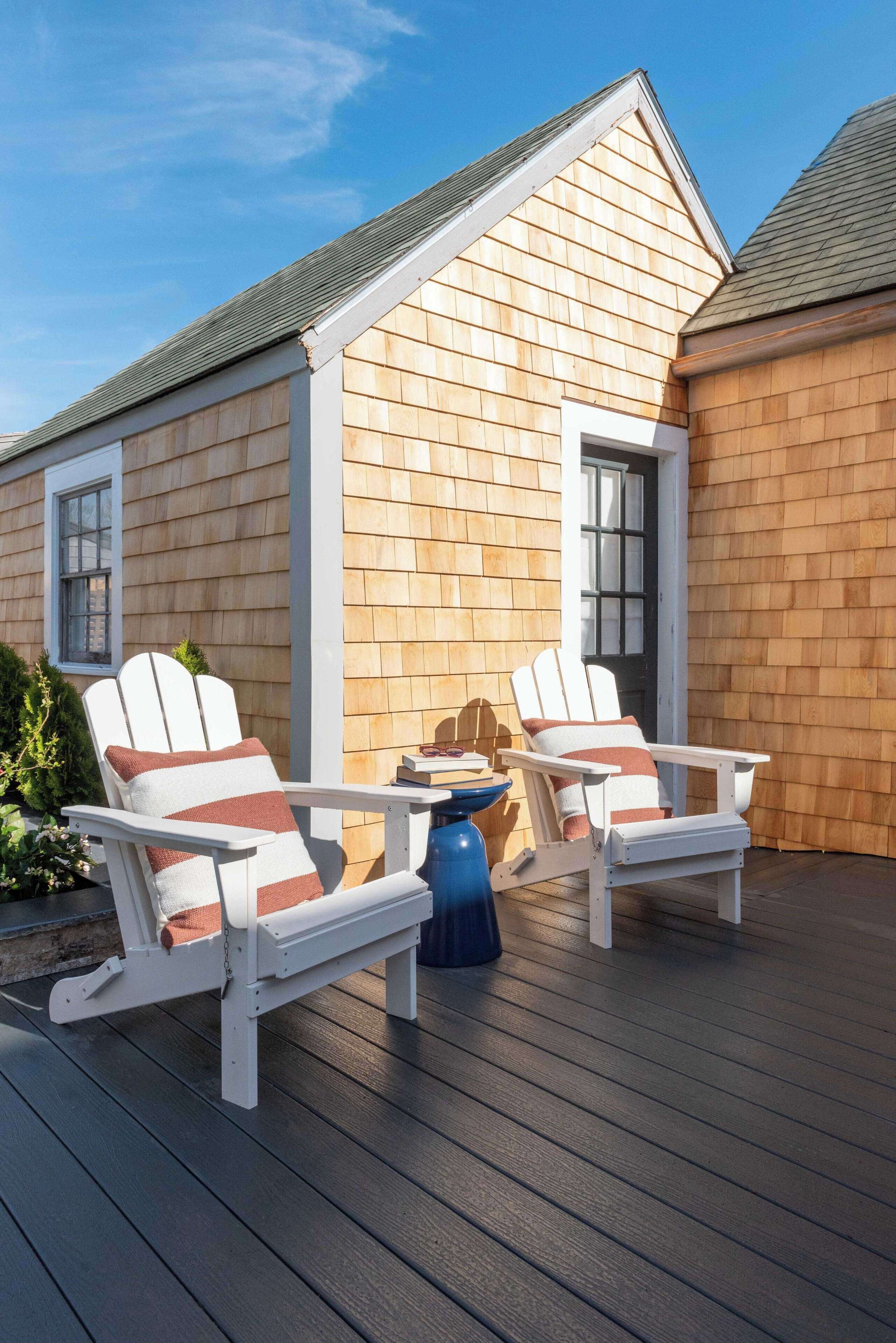 A deck with two Adirondack chairs and a small table, set against a wooden house with a gabled roof.