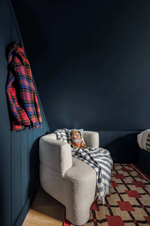 A corner room with a dark blue wall, a plaid jacket hanging on the wall, a white armchair with a striped blanket, and a patterned rug. - Beautiful Historic Home, Heated Pool & Hot Tub (Nantucket)