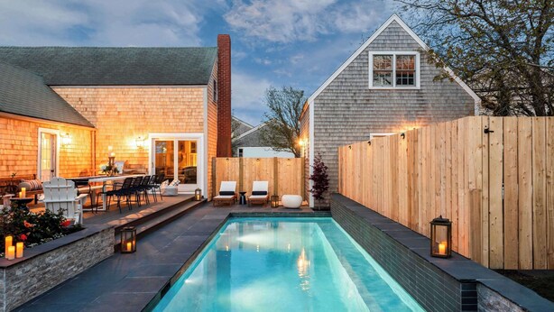 A rectangular swimming pool with a wooden deck and lounge chairs, surrounded by a wooden fence and a brick wall. - Beautiful Historic Home | Heated Pool & Hot Tub (Nantucket)