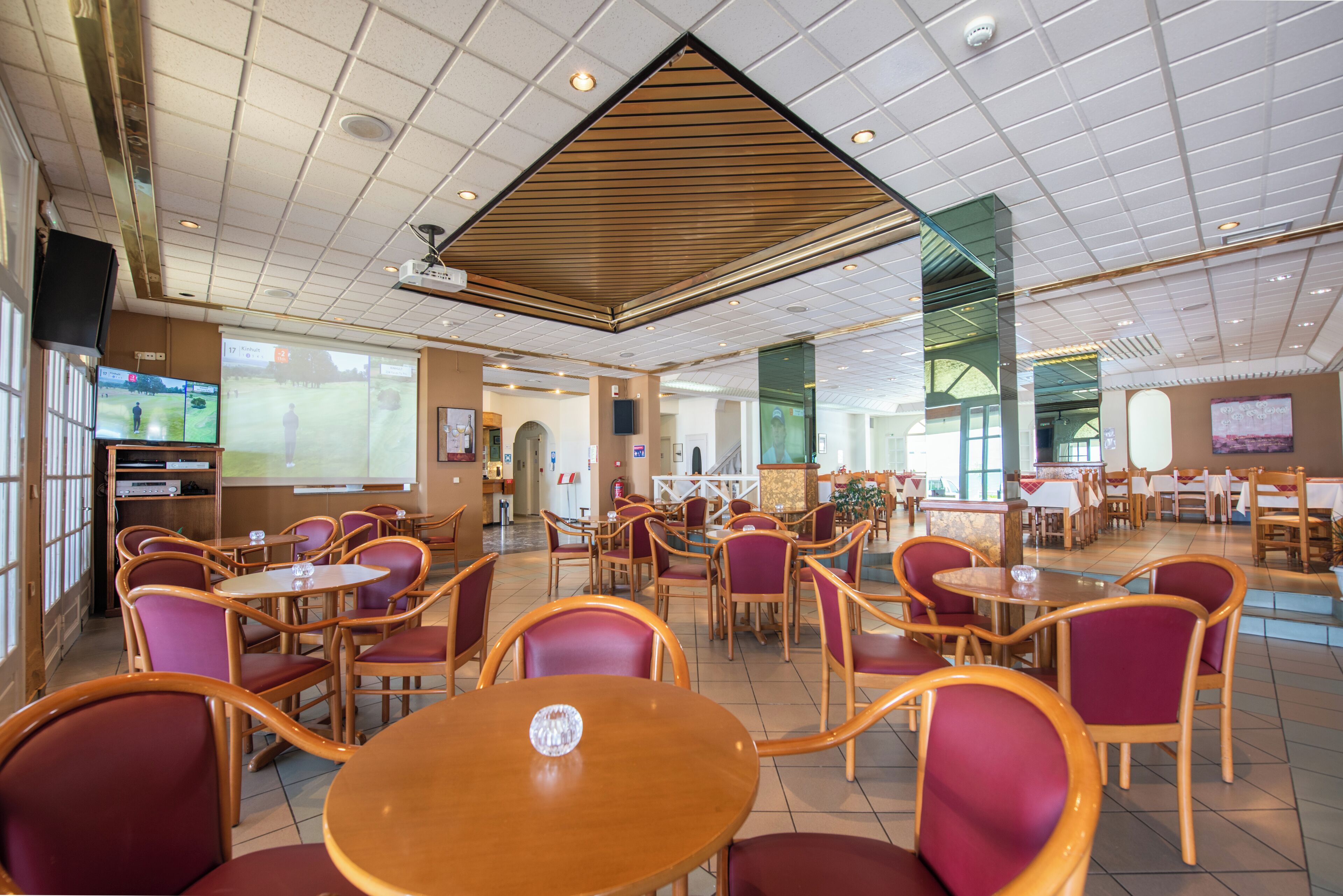 A spacious restaurant with multiple tables and chairs, a large screen displaying a golf game, and a projector screen in the center.