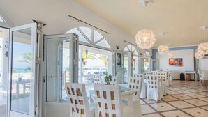 A dining area with white chairs and tables, large windows, and decorative hanging lights. - Aphrodite Beach Hotel Corfu (Corfu)