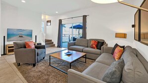 Una sala de estar moderna con sofá, sillón y mesa de centro, televisor de pantalla plana y un amplio ventanal con cortinas.