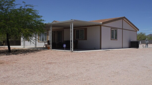 A single-story building with a covered patio area, a gravel driveway, and a tree to the left.