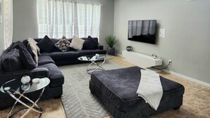 Un salon moderne avec un canapé d’angle, une table basse en verre, un téléviseur à écran plat fixé au mur et une plante en pot.