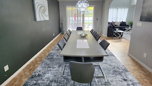 Une salle à manger moderne avec une longue table, des chaises, un lustre et une vue sur un salon comprenant un canapé et une table basse.
