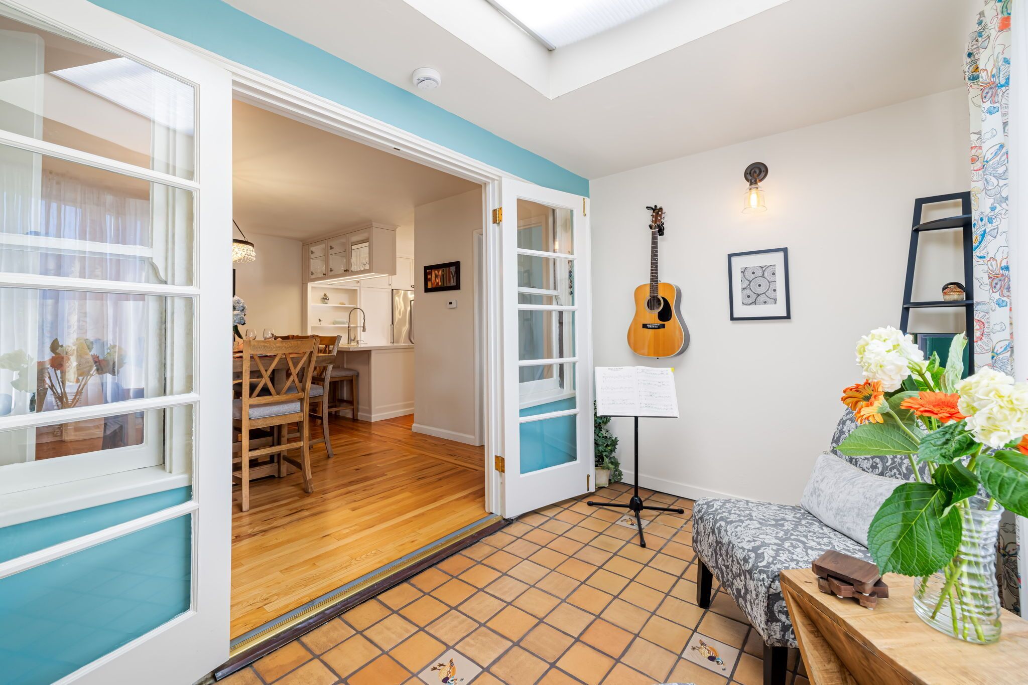 A cozy living room with a wooden floor, a dining area with a table and chairs, a guitar, and a vase with flowers.