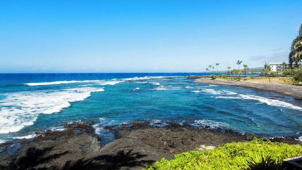 A coastal view with clear blue waters, rocky shoreline, and a distant building with palm trees. - Kksr#48 Beautiful Townhome, Steps from the Pool/Beach, Remodeled, AC in unit! (KAILUA KONA)