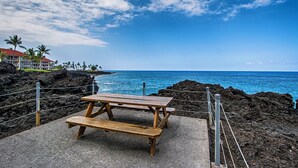 A wooden picnic table with benches on a concrete platform, overlooking a rocky coastline with the ocean and a clear sky. - Kksr#48 Beautiful Townhome, Steps from the Pool/Beach, Remodeled, AC in unit! (KAILUA KONA)