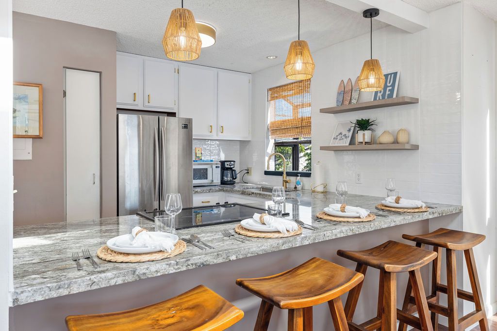 A modern kitchen with a marble countertop, stainless steel appliances, wooden stools, and pendant lighting.
