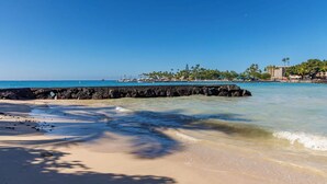 A beach with clear blue water, a rocky breakwater, and a line of palm trees and buildings in the background.