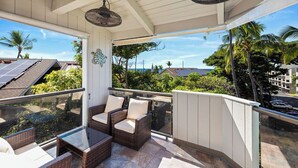 A covered outdoor seating area with wicker furniture and a glass balustrade, overlooking a residential neighborhood with palm trees and solar panels on rooftops.