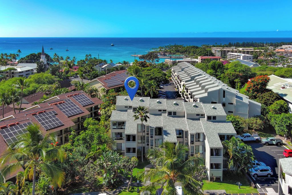 A multi-story building with a rooftop garden and solar panels, surrounded by lush greenery and a view of the ocean.