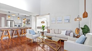 A modern living room with a sofa, coffee table, and wooden stools. A kitchen with a refrigerator and stove is visible in the background.