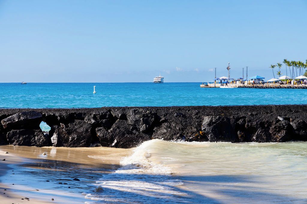 A beach with a rocky breakwater, clear blue water, and a distant cruise ship.