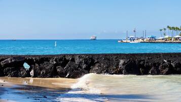 A beach with a rocky breakwater, clear blue water, and a distant cruise ship.