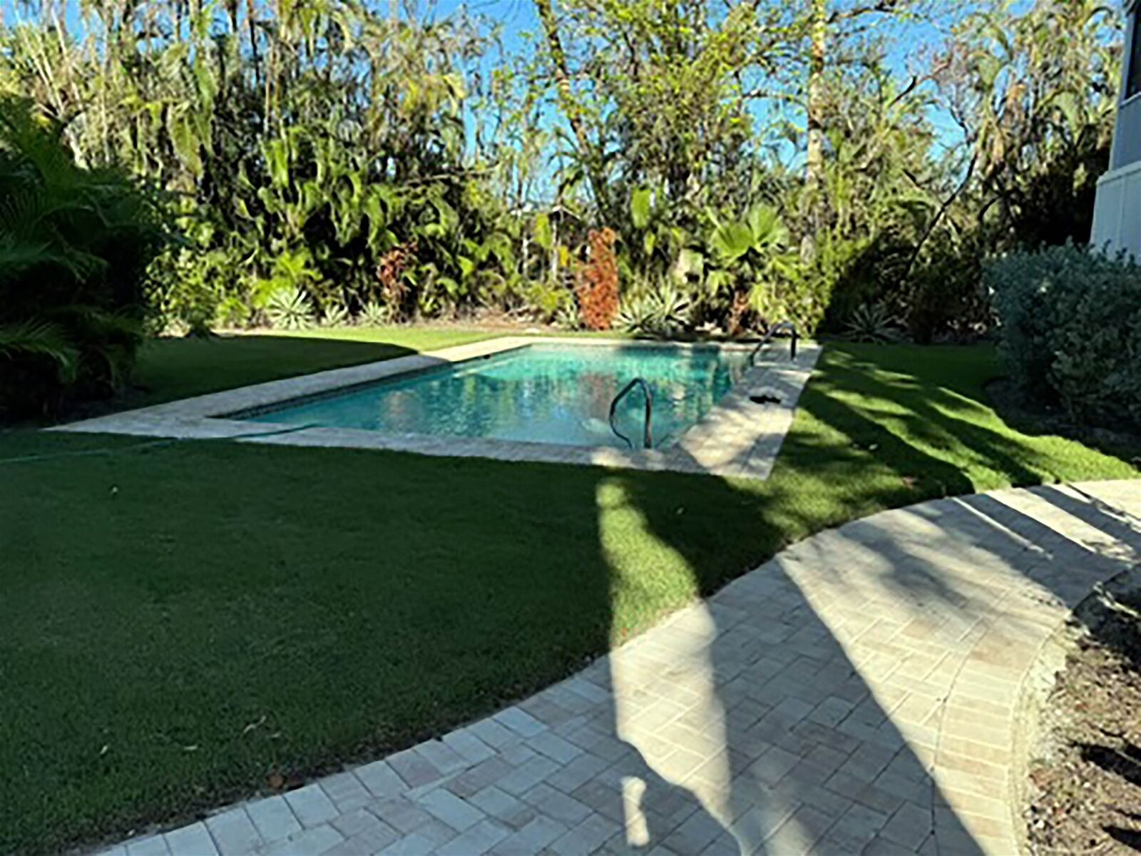 Une piscine rectangulaire dotée d’une terrasse dallée, entourée d’une pelouse bien entretenue et de diverses plantes.