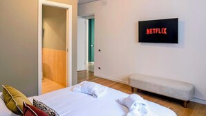 A modern hotel room with a bed, a bench, a TV displaying the Netflix logo, and a door leading to another room. - Exclusive Apartment with Jacuzzi in the Historic Center of Palermo (Palermo)