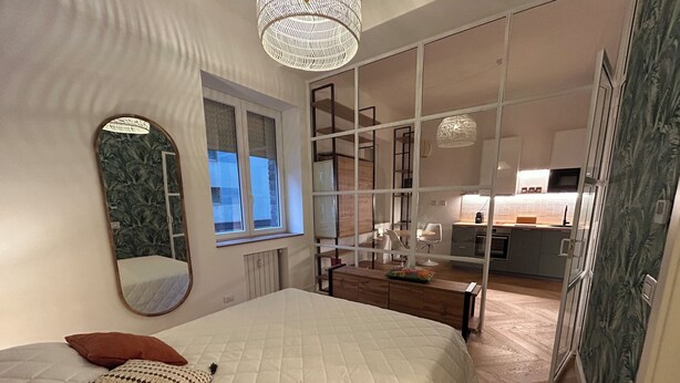 A bedroom with a large bed, a bedside mirror, and a view of the kitchen through a glass partition. - Comfort chic - Duomo in 5 minutes (Milano)