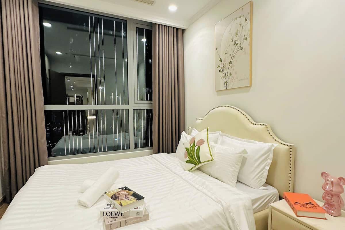 Una cama bien hecha con una colcha blanca y almohadas, una mesita de noche con un libro y un adorno, y una ventana con cortinas.