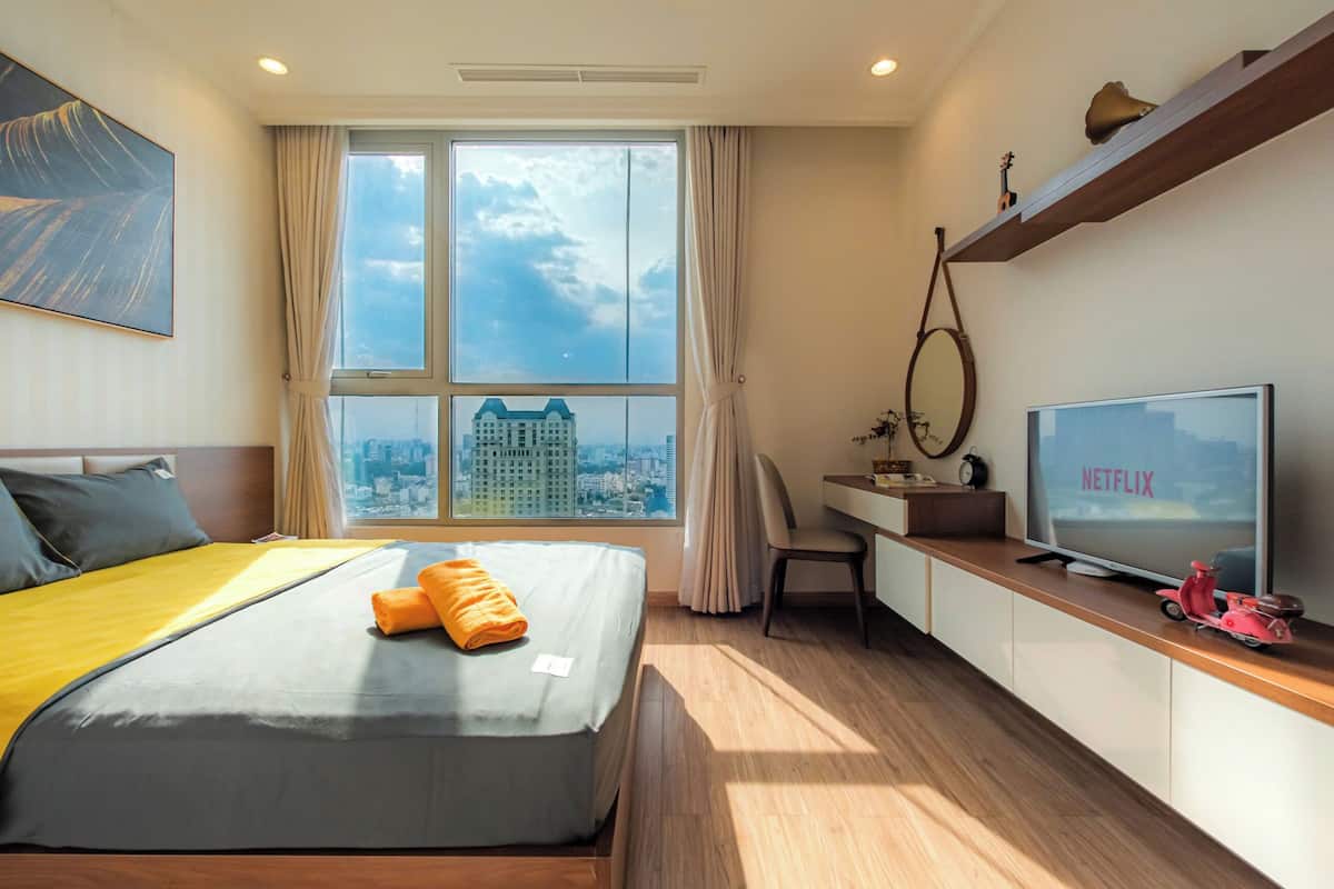 Habitación de hotel con cama, escritorio, televisor mostrando 'NETFLIX' y vistas de un paisaje urbano a través de amplias ventanas.