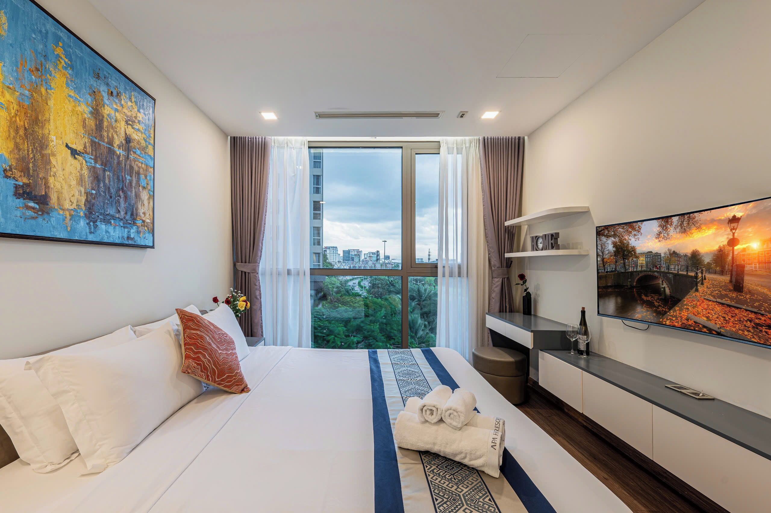 A hotel room with a large bed, a television, a view of the city, and a painting on the wall.