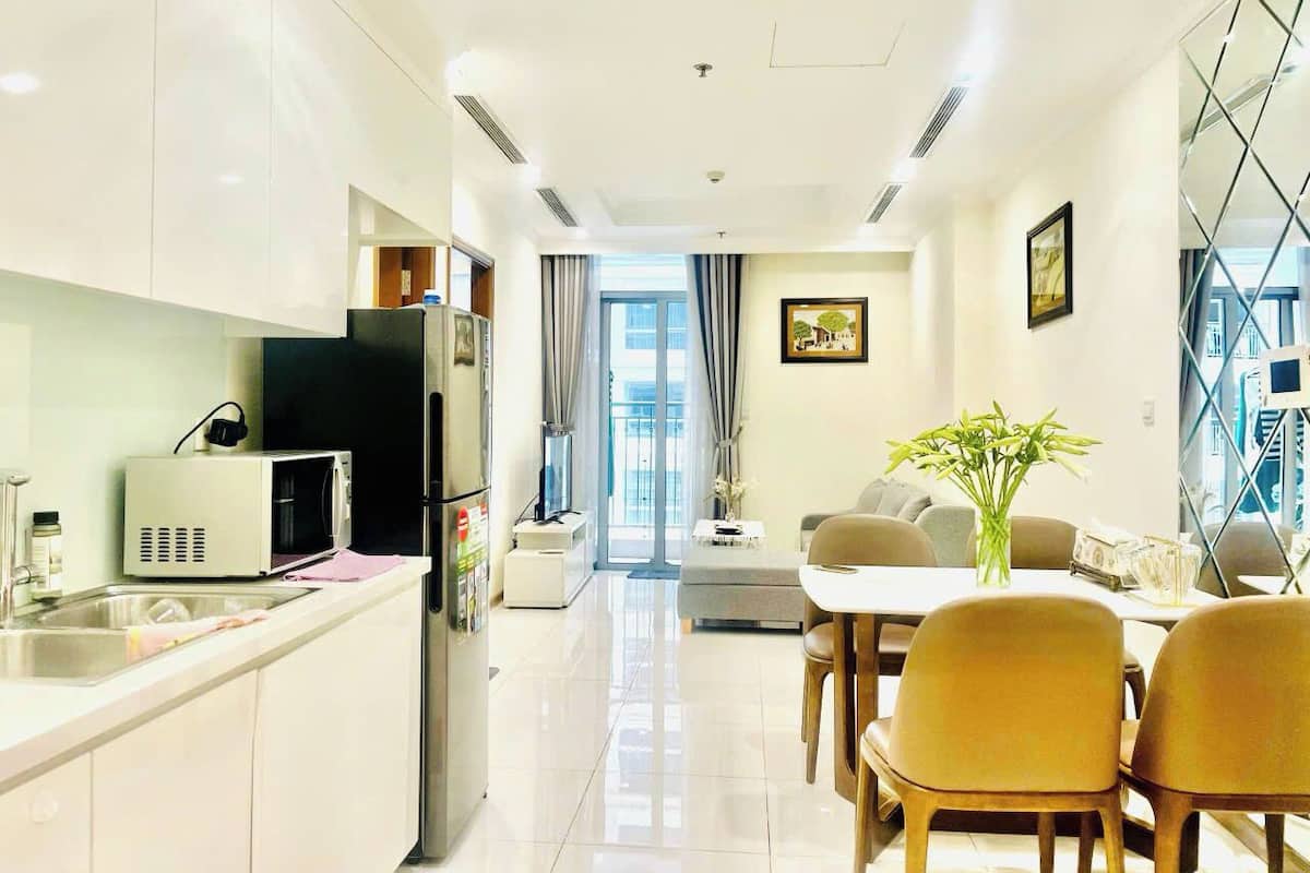 Una cocina moderna con armarios blancos, microondas y un lavamanos. Hay una zona de comedor con una mesa y sillas, y un sofá.